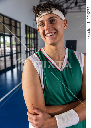 Teenage male basketball player in green jersey standing with crossed arms on court near windows 141542754