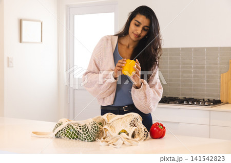 Asian woman holding yellow bell pepper and inspecting mesh bags and red pepper on island countertop Asian woman holding yellow bell pepper and inspecting mesh bags and red pepper on island countertop 141542823