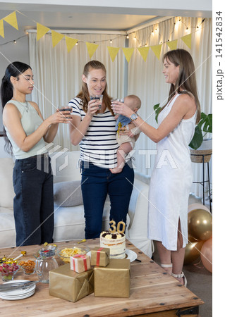 Diverse female friends clinking glasses with baby in living room with birthday cake, balloons Diverse female friends clinking glasses with baby in living room with birthday cake, balloons 141542834