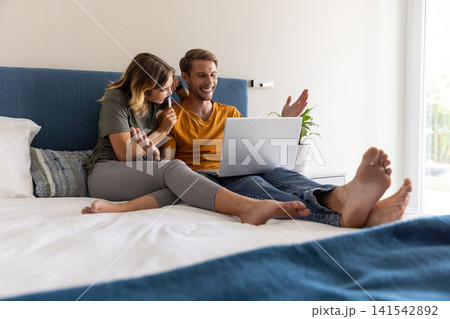 Couple working on laptop sitting on bed in bedroom and holding pen near potted plant 141542892