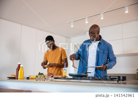 African american father and son cooking on kitchen island with spatula, stainless steel frying pan African american father and son cooking on kitchen island with spatula, stainless steel frying pan 141542961