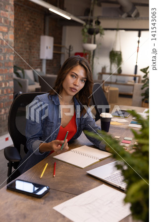Woman wearing denim shirt over red top holding pencil above notebook at coworking desk 141543063