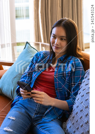 Asian woman holding smartphone on brown sofa in living room with glass side table Asian woman holding smartphone on brown sofa in living room with glass side table 141543124