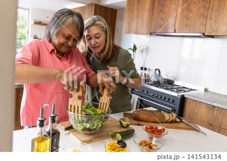 Diverse senior friends tossing salad at kitchen island with bowl, wooden servers, vegetables Diverse senior friends tossing salad at kitchen island with bowl, wooden servers, vegetables 141543134
