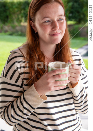 Female with red hair wearing beige sweater holding green mug on garden patio in sunlight 141543135
