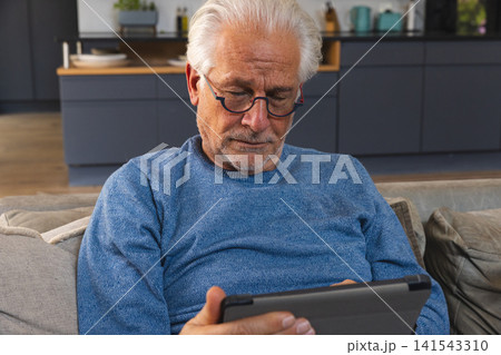 Senior man wearing blue sweater sitting on sofa with eyeglasses and using tablet in kitchen lounge 141543310