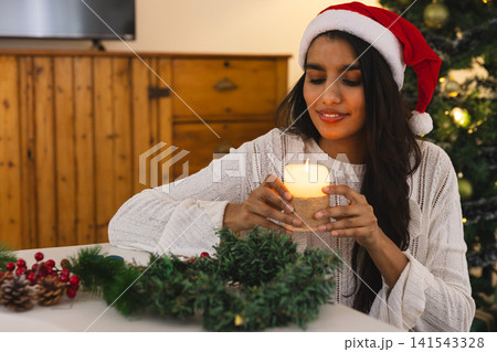 Asian woman sitting in living room making wreath holding lit candle wearing sweater and Santa hat 141543328