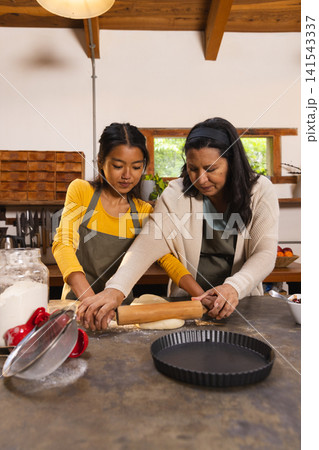 Diverse mother and daughter in aprons rolling dough with rolling pin on countertop in home kitchen Diverse mother and daughter in aprons rolling dough with rolling pin on countertop in home kitchen 141543337