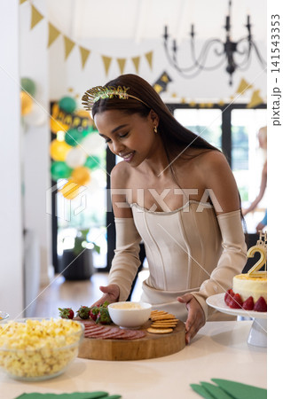 African american woman wearing strapless dress arranging buffet at home with charcuterie, cake 141543353