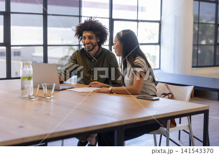 Diverse coworkers leaning forward and smiling while reviewing papers on laptop in office Diverse coworkers leaning forward and smiling while reviewing papers on laptop in office 141543370
