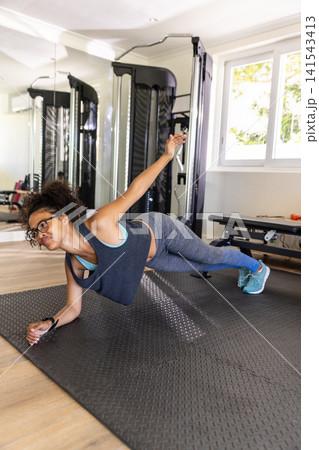 African American woman performing side plank on black textured mat in home gym with cable machine African American woman performing side plank on black textured mat in home gym with cable machine 141543413