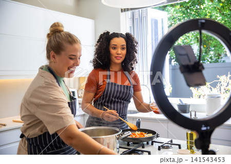 Diverse female friends stirring, cooking chopped vegetables on kitchen island with ring light 141543453