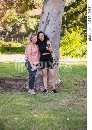 Diverse female friends leaning against tree at park taking selfie with phone, water bottle Diverse female friends leaning against tree at park taking selfie with phone, water bottle 141543454