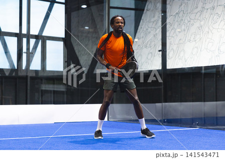 African American man wearing sportswear standing on padel court holding padel racket in glass arena African American man wearing sportswear standing on padel court holding padel racket in glass arena 141543471