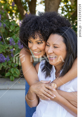 Diverse mother and daughter hugging in backyard patio with planter, blossoms and wristwatch Diverse mother and daughter hugging in backyard patio with planter, blossoms and wristwatch 141543554