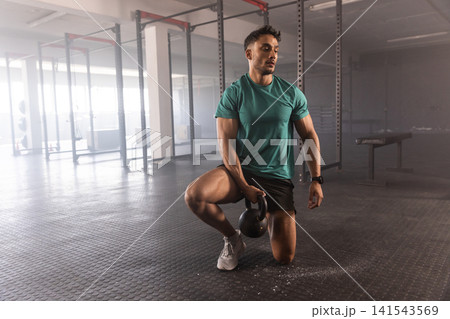 Kneeling man wearing teal shirt gripping black kettlebell on gym floor near squat rack, copy space Kneeling man wearing teal shirt gripping black kettlebell on gym floor near squat rack, copy space 141543569