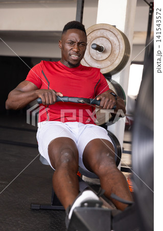 African American male athlete in red shirt pulling rowing handle at fitness gym near weight plates 141543572