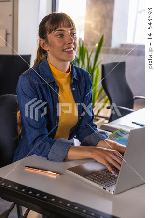 Mid adult woman wearing denim shirt typing on laptop at office desk with metallic pen Mid adult woman wearing denim shirt typing on laptop at office desk with metallic pen 141543593