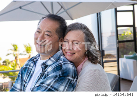 Diverse couple hugging on terrace loveseat under patio umbrella overlooking garden Diverse couple hugging on terrace loveseat under patio umbrella overlooking garden 141543641