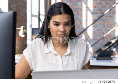 Woman typing on laptop at wooden desk in office loft with monitor and brick wall Woman typing on laptop at wooden desk in office loft with monitor and brick wall 141543675