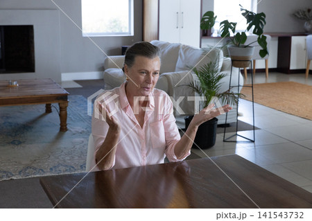 Senior woman gesturing with hands while talking at dining table in living room next to plants Senior woman gesturing with hands while talking at dining table in living room next to plants 141543732