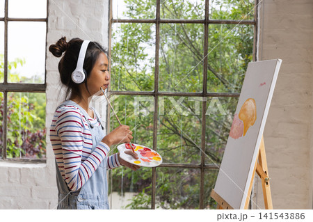 Asian woman standing in art studio holding palette and paintbrush between lips in front of canvas Asian woman standing in art studio holding palette and paintbrush between lips in front of canvas 141543886