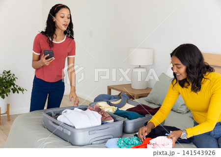 Diverse mom and teenage daughter packing clothes into open grey suitcase on bed Diverse mom and teenage daughter packing clothes into open grey suitcase on bed 141543926