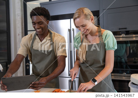 Diverse couple prepping meal at kitchen island using tablet, slicing carrots on cutting board 141543967
