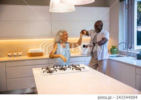 Diverse senior couple dancing hand in hand in modern kitchen near labeled canisters and gas cooktop Diverse senior couple dancing hand in hand in modern kitchen near labeled canisters and gas cooktop 141544004
