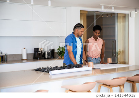 Diverse couple standing at kitchen island by gas cooktop examining blueprints holding coffee mugs 141544018