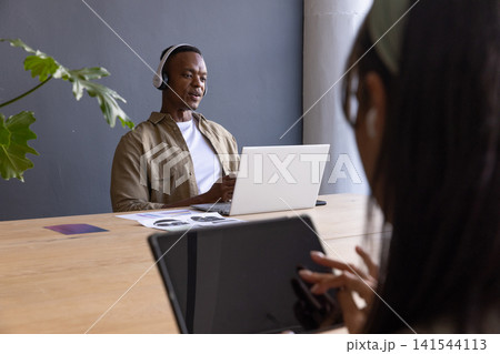 African american coworkers using laptop, tablet, headphones at table with plant, copy space 141544113