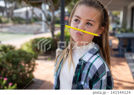 Girl balancing pencil under nose outdoors on sunlit brick walkway near patio seating 141544124
