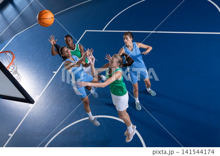 Diverse female opponents jumping and reaching for orange basketball under hoop on blue court Diverse female opponents jumping and reaching for orange basketball under hoop on blue court 141544174