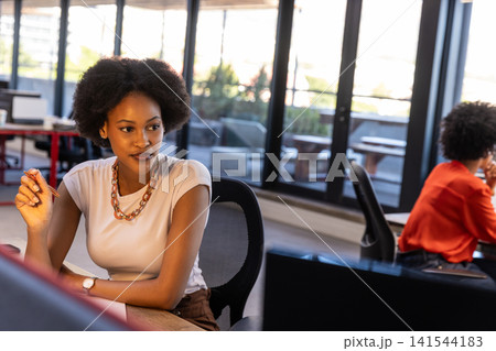 Diverse female coworkers sitting on ergonomic chairs at desk in office holding pen by monitors Diverse female coworkers sitting on ergonomic chairs at desk in office holding pen by monitors 141544183