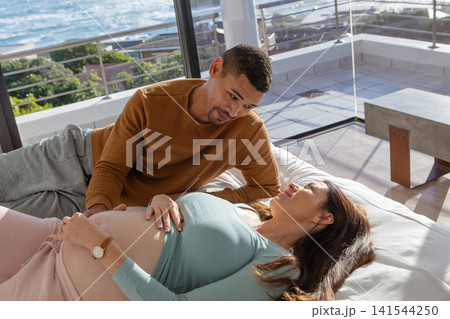 Diverse couple leaning on bed caressing pregnant belly in bedroom with ocean view by glass doors Diverse couple leaning on bed caressing pregnant belly in bedroom with ocean view by glass doors 141544250