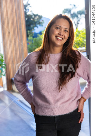 Mature asian woman standing at patio door wearing pink sweater with hands on hips smiling at camera Mature asian woman standing at patio door wearing pink sweater with hands on hips smiling at camera 141544259