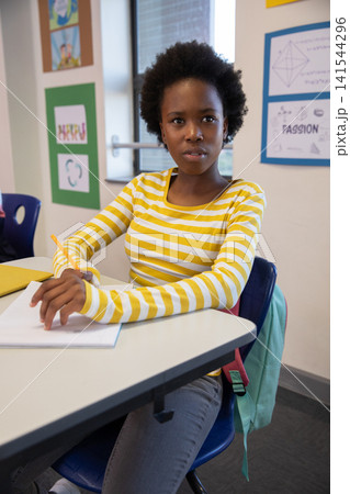 African american girl student writing in notebook at school desk with pencil, backpack, posters African american girl student writing in notebook at school desk with pencil, backpack, posters 141544296