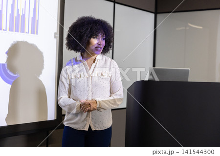 Mid adult woman in blouse presenting bar and pie charts on screen at conference podium 141544300