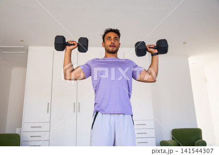 Asian man lifting black hex dumbbells at shoulder height in home gym in purple sportswear 141544307