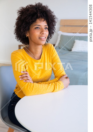 African american woman sitting at table in bedroom wearing yellow top, gazing African american woman sitting at table in bedroom wearing yellow top, gazing 141544308