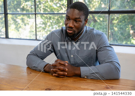 African american man sitting at table in meeting room, clasping hands near window African american man sitting at table in meeting room, clasping hands near window 141544330