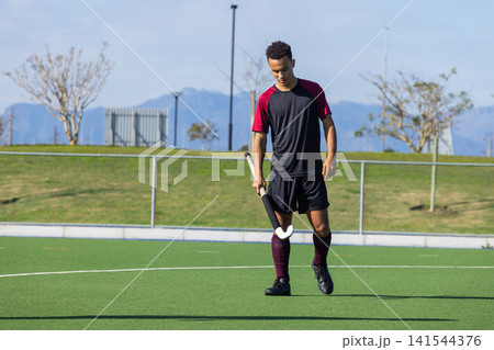 African american male athlete walking on turf field with hockey stick near metal fence African american male athlete walking on turf field with hockey stick near metal fence 141544376
