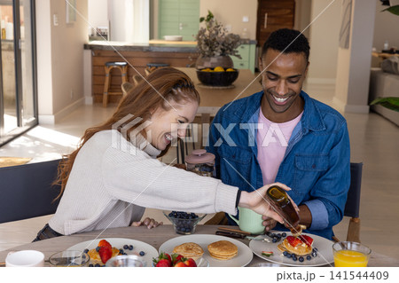 Diverse couple pouring syrup onto pancakes at dining table at home with coffee press Diverse couple pouring syrup onto pancakes at dining table at home with coffee press 141544409