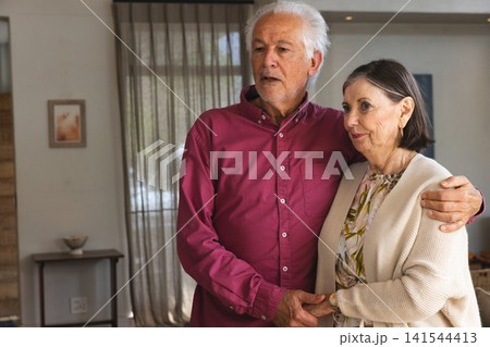 Senior couple standing in living room by window with sheer curtains near side table with bowl Senior couple standing in living room by window with sheer curtains near side table with bowl 141544413