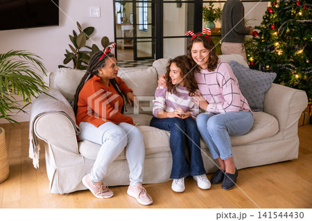 Diverse family chatting, sitting on sofa in living room with christmas tree, antler headbands 141544430