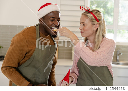Diverse couple wearing green aprons and red party hat feeding each other cookies at modern kitchen Diverse couple wearing green aprons and red party hat feeding each other cookies at modern kitchen 141544438