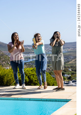 Diverse female friends standing on patio by poolside shrubs and popping golden confetti Diverse female friends standing on patio by poolside shrubs and popping golden confetti 141544449