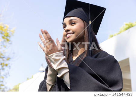 Female graduate smiling and clapping in black mortarboard and gown at graduation Female graduate smiling and clapping in black mortarboard and gown at graduation 141544453