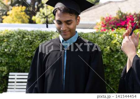 Asian man in graduation gown, mortarboard, examining pen being offered by classmate in backyard 141544454
