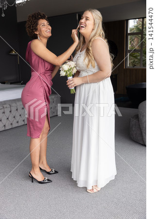 Diverse bride and bridesmaid sporting gowns laughing holding bouquet, smartphone, in bridal suite Diverse bride and bridesmaid sporting gowns laughing holding bouquet, smartphone, in bridal suite 141544470